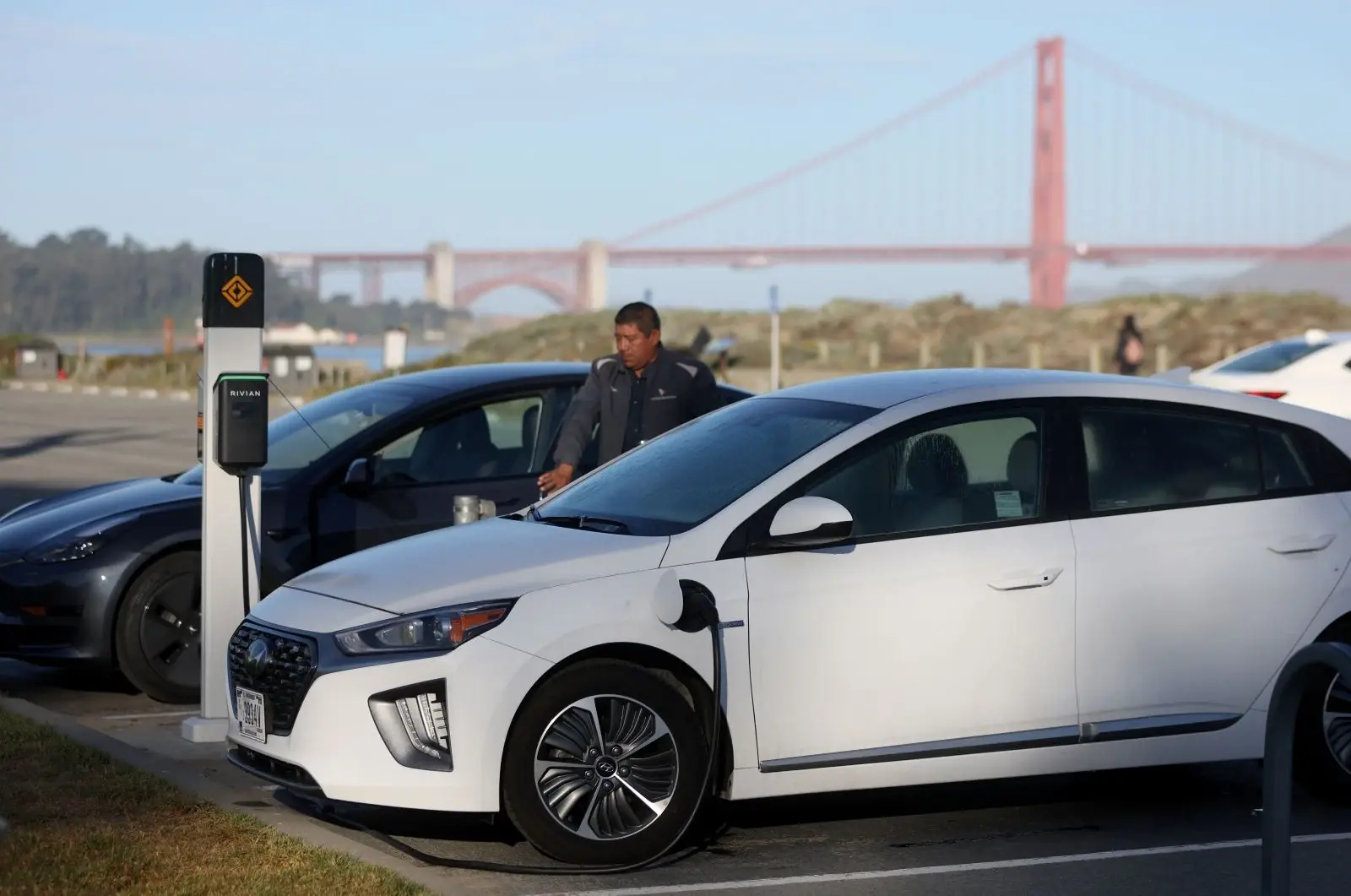 A man exits his electric vehicle at a charging station, with the Golden Gate Bridge visible in the distance
