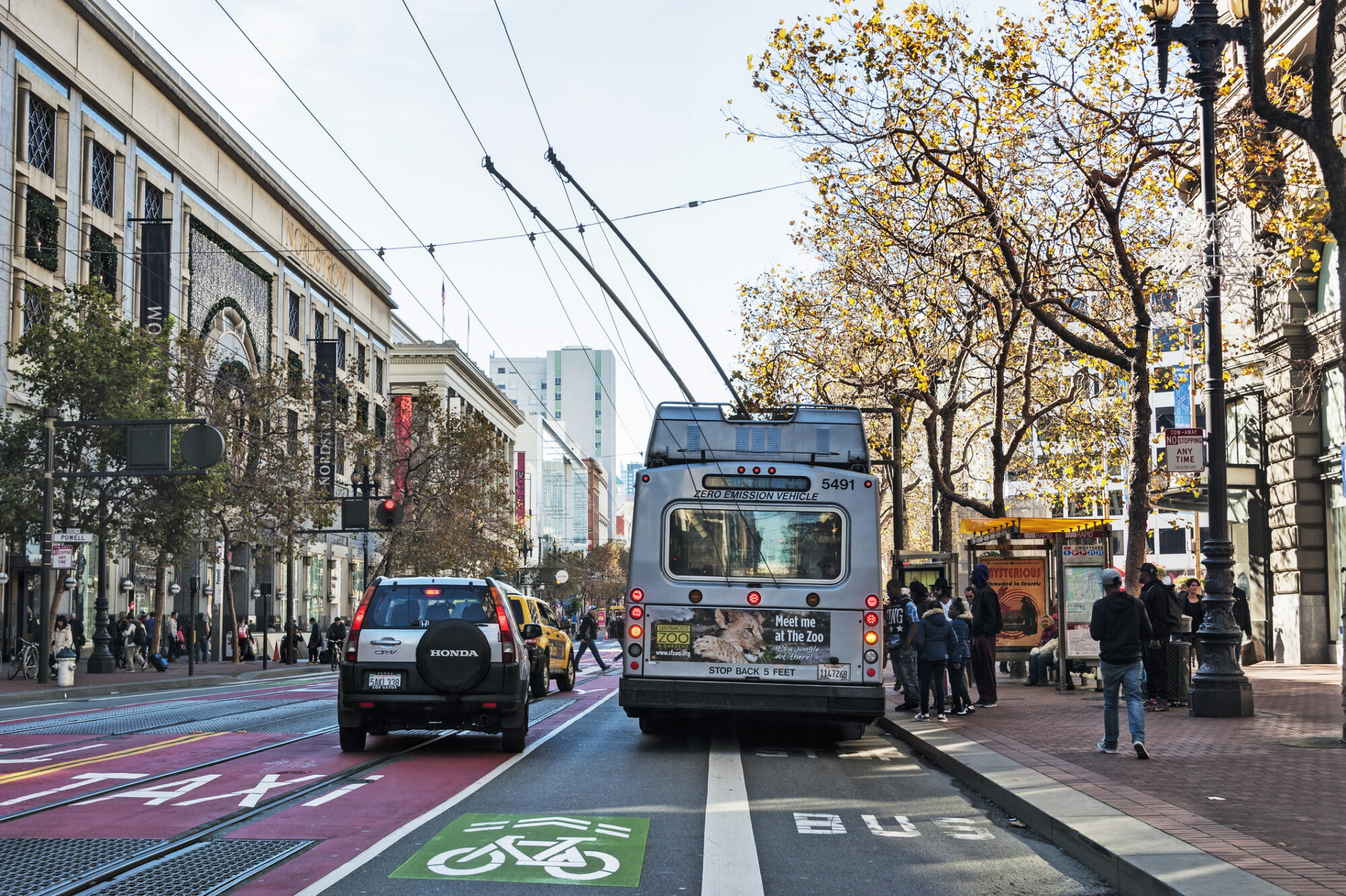 An electric bus on a busy San Francisco street loads passengers while cars and pedestrians are passing by. Three lanes are visible on the street: from left to right, the first displays the word 