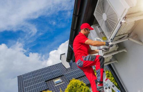 An HVAC technician dressed in red on a ladder beside a home performing maintenance work on two heat pump units. Another home appears in the background with solar panels installed on the roof.
