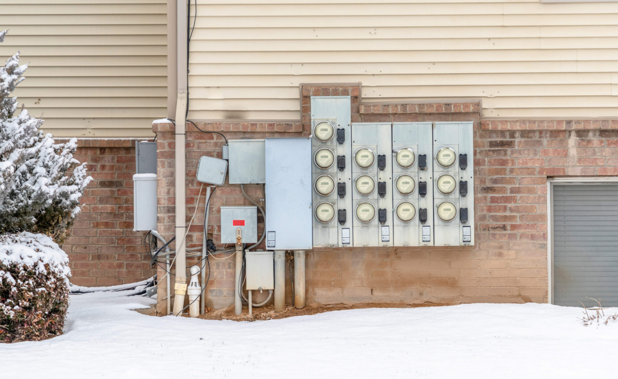 Exterior of residential building with multiple electricity meters mounted on brick wall. The surrounding yard is blanketed with snow.