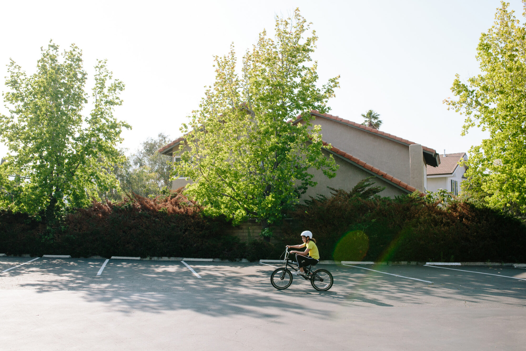 A child wearing a helmet and yellow shirt rides a BMX bike across an empty parking lot, with trees and a house in the background.