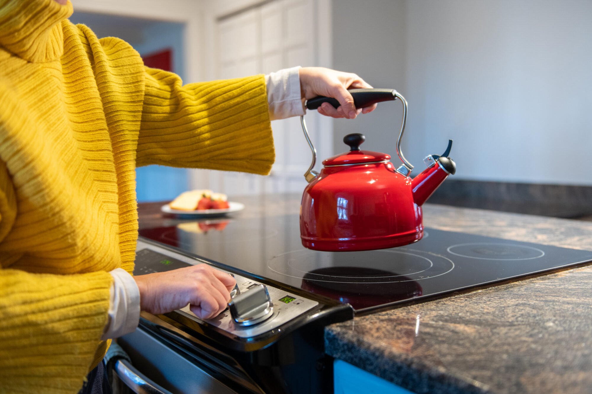 A person in a yellow sweater places a red kettle on an electric glass-top stove while adjusting the control knob.
