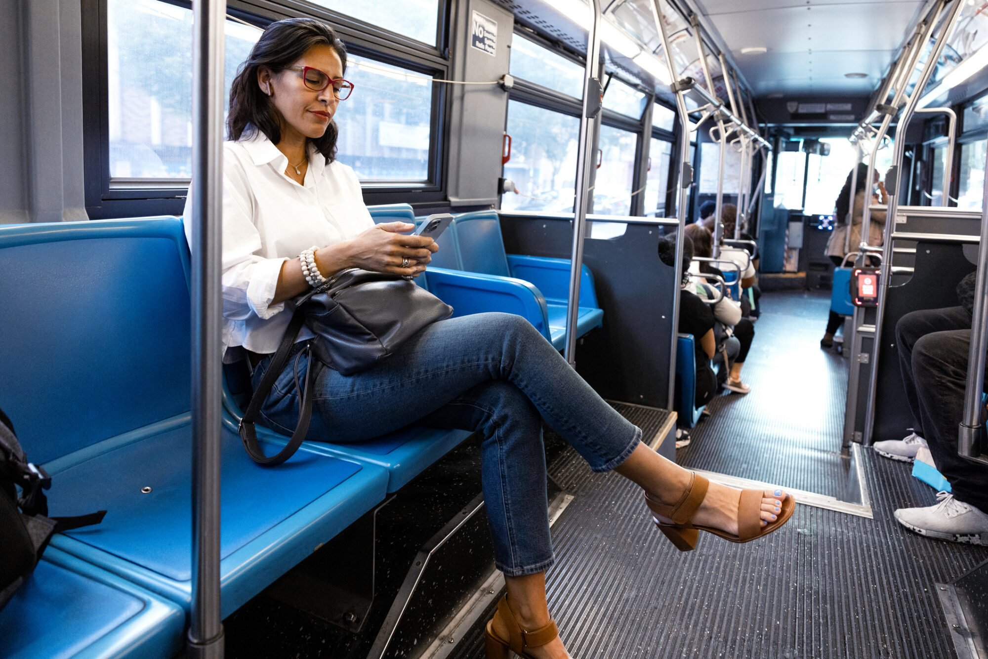 A woman sits on a public bus looking at her phone, with other passengers seated further back.