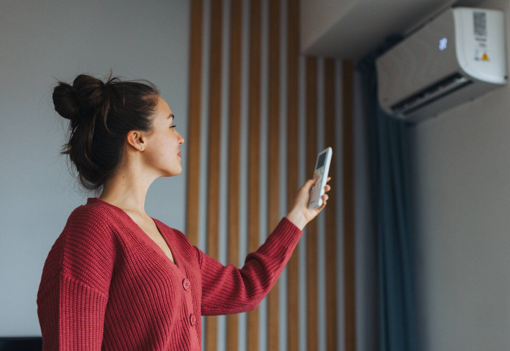 A woman in a red sweater points a remote control at a wall-mounted air conditioning/heating unit.
