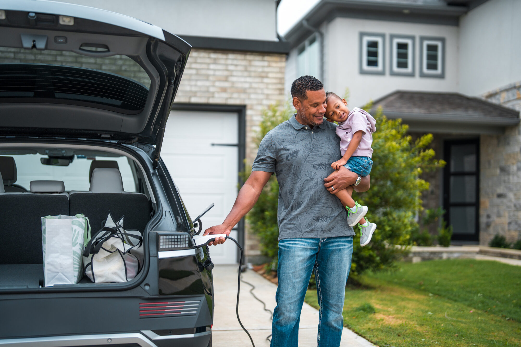A man holds a young child while charging his electric vehicle in the driveway of a home, with the trunk open and grocery bags visible inside.