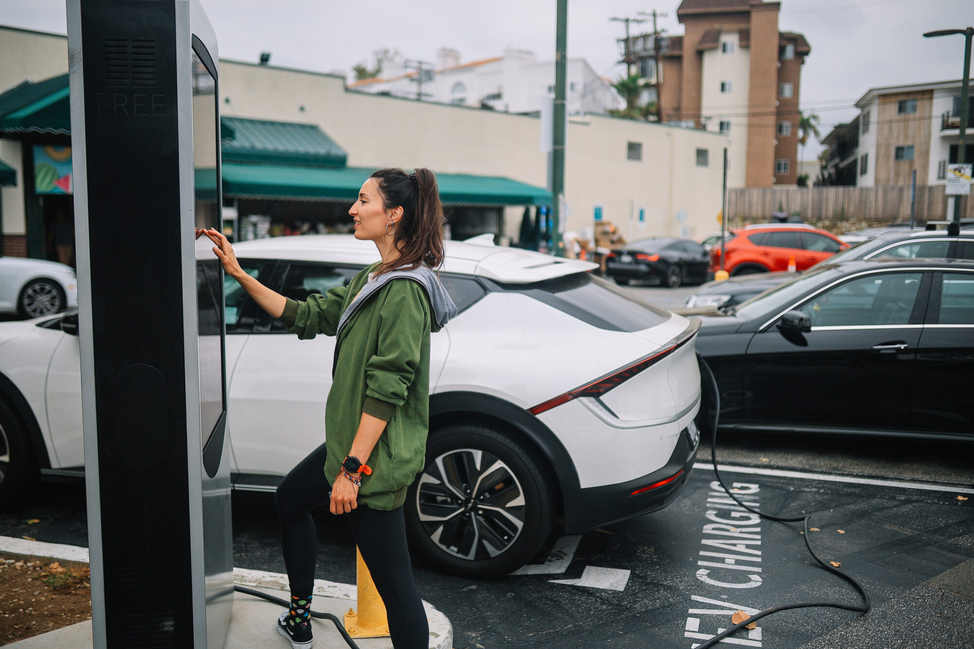 A woman uses a public EV charging station in a parking lot in front of a strip mall to charge her white electric car, parked in a designated 'EV Charging' spot.
