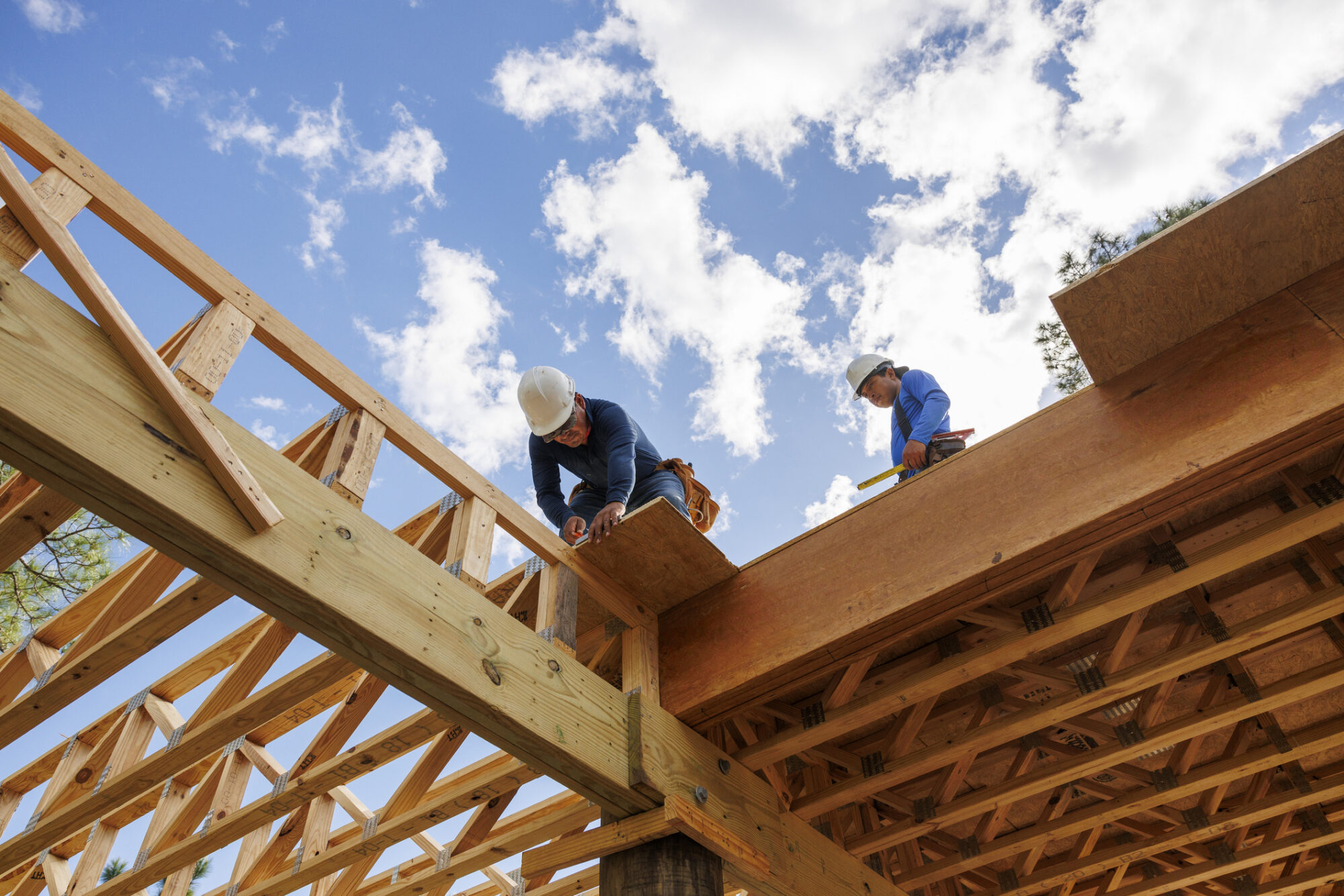 A low-angle view of two construction workers in hard hats working on the wooden roof trusses of a house under construction, against a blue sky with scattered clouds.