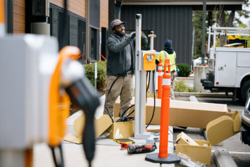 Workers install a new EV charging station outdoors in a parking area, surrounded by tools, cardboard packaging, and traffic cones.