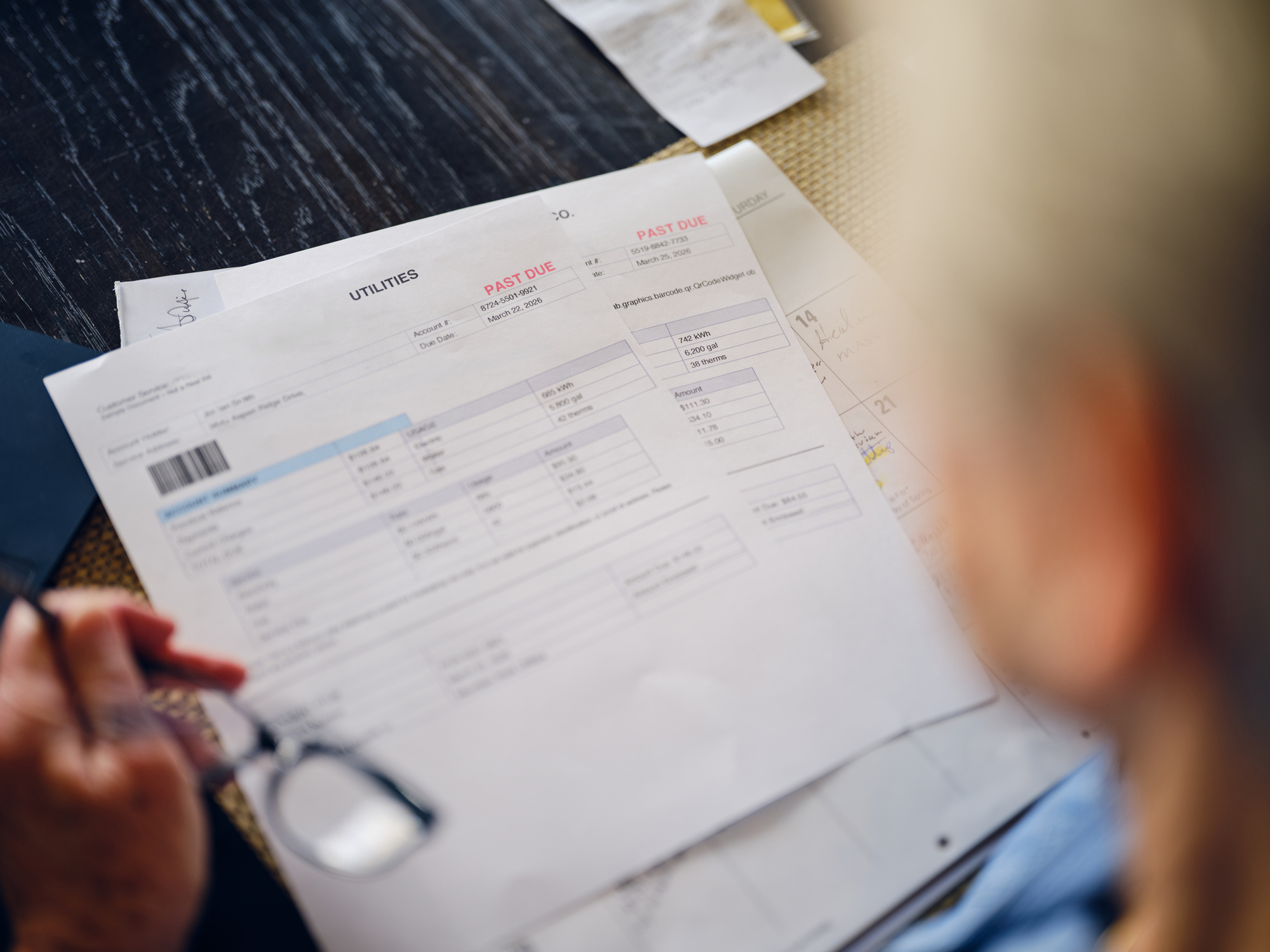 A person holds a pair of reading glasses while reviewing overdue utility bills marked 'PAST DUE,' as seen from their vantage point.