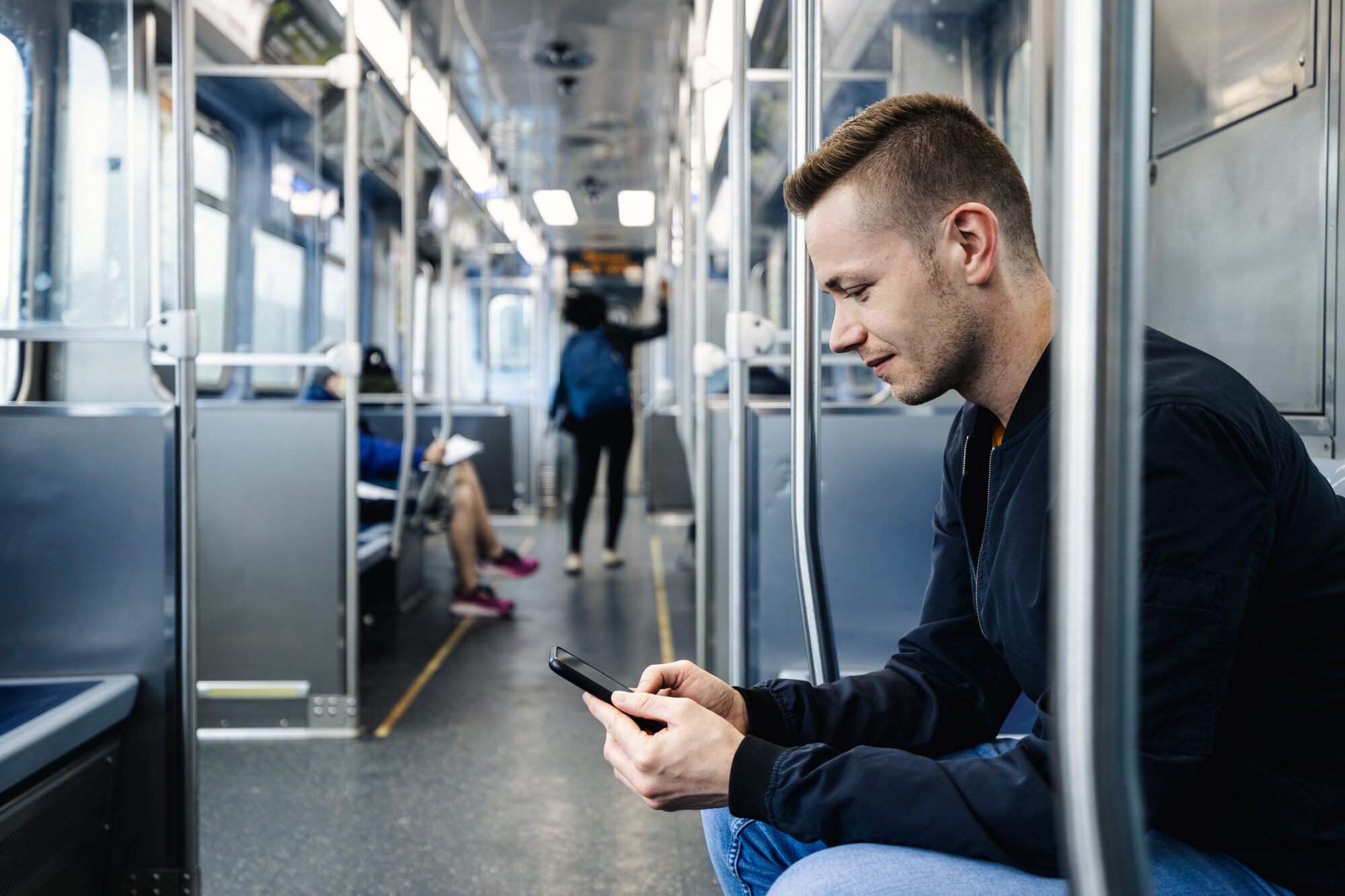 A man sits looking at his phone while riding a subway or train car, with other passengers visible in the background.