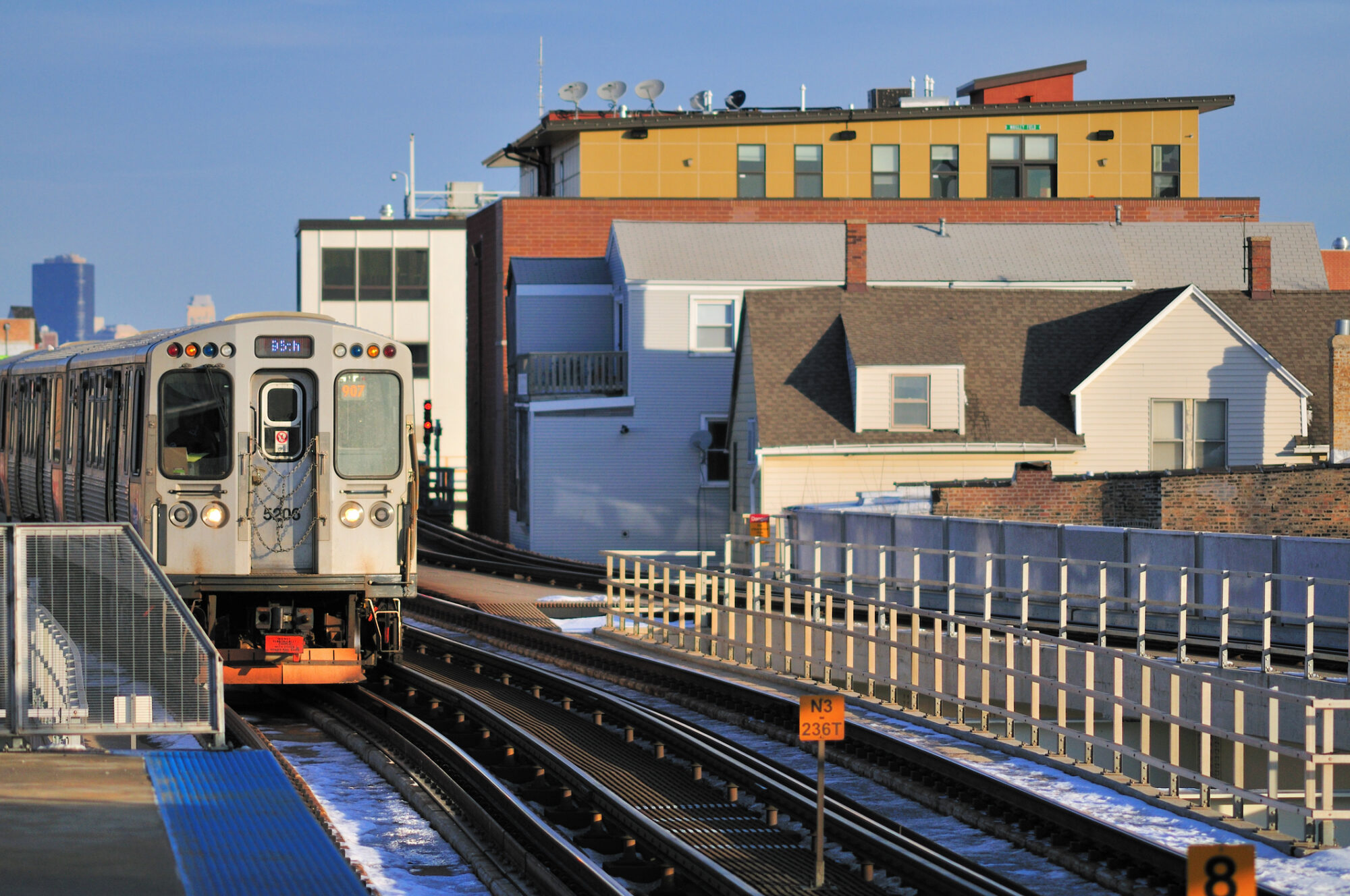 An elevated train approaches a station platform along a curved track, with residential and commercial buildings in the background on a clear day.