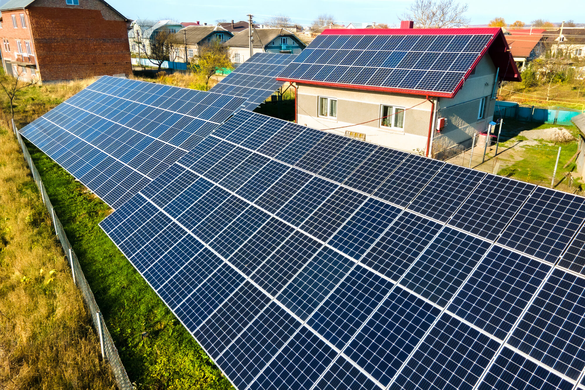 An aerial view of a large ground-mounted solar panel array beside a house with a red roof, in a residential neighborhood.