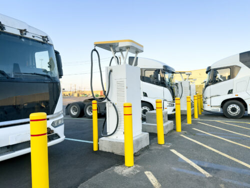 An electric charging station stands between rows of parked white semi trucks, with a thick charging cable connected to one of the vehicles in the fleet parking lot. Yellow safety bollards surround the charging unit, protecting it from vehicle contact within the paved lot. Painted parking lines extend across the asphalt, with power lines and distant buildings visible along the horizon under a clear sky.