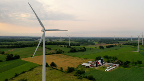 An aerial view of a wind turbine in Wisconsin towering over farmland, with a red barn and silos below and more turbines dotting the rolling green fields in the distance.