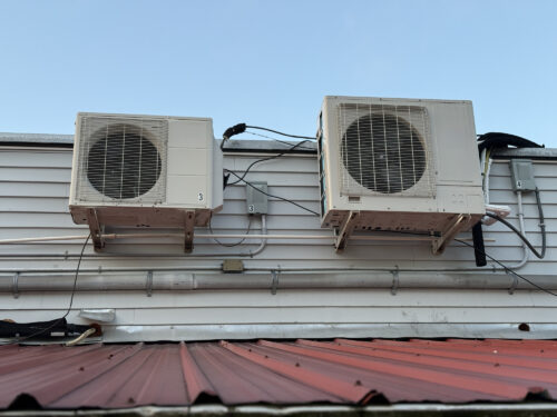 Two outdoor mini-split air conditioning/heat pump condenser units mounted on brackets on the exterior wall of a building, above a red metal roof